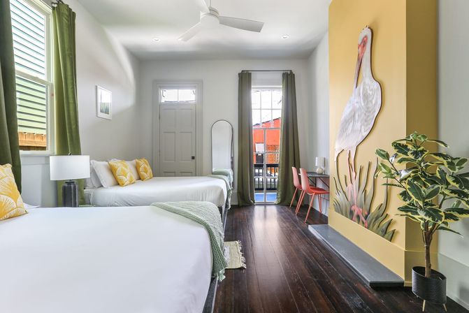 Bright coastal-inspired guest room with two white beds, yellow accent pillows, green curtains, dark hardwood floors, balcony door, desk with pink chairs, and a large playful heron mural on a yellow accent wall.