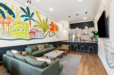 Open-concept apartment interior with green sofas, coffee table and rug, vibrant tropical cityscape mural on the wall, breakfast bar with metal stools and pendant lights, stainless-steel kitchen and wood-look flooring.