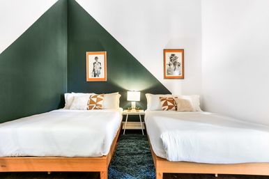 Modern guest bedroom with two twin wooden beds and white linens, a dark green triangular accent wall, central nightstand with lamp and succulents, geometric throw pillows and framed black-and-white art
