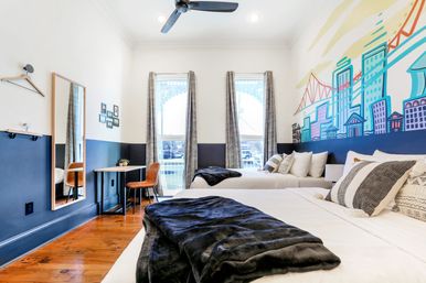 Bright modern guest bedroom with two beds, navy half-wall and colorful city skyline mural, hardwood floors, two tall windows with patterned curtains, small desk with leather chair, wall mirror and ceiling fan.