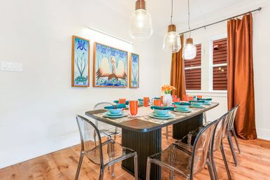 Bright contemporary home dining room with a dark wood table set for eight featuring teal plates and bowls, orange glassware, clear acrylic chairs, hanging glass pendant lights, rust-colored curtains, and a three-panel swan artwork on the wall.