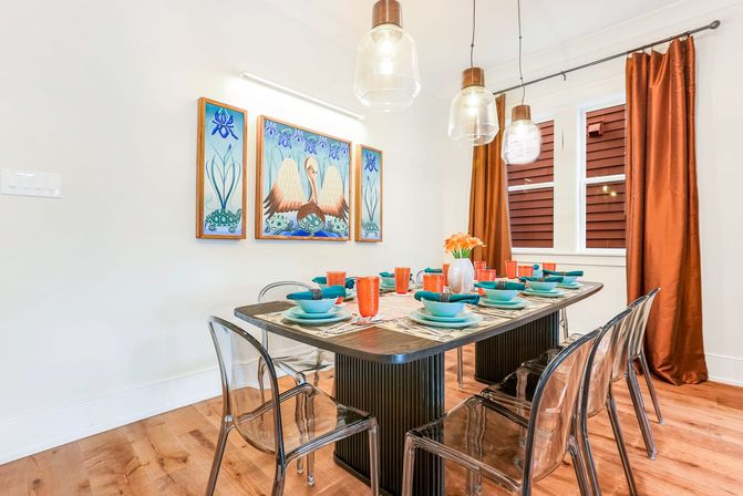 Bright contemporary home dining room with a dark wood table set for eight featuring teal plates and bowls, orange glassware, clear acrylic chairs, hanging glass pendant lights, rust-colored curtains, and a three-panel swan artwork on the wall.