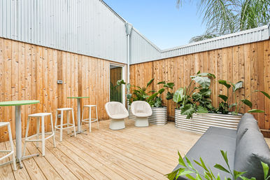 Bright modern outdoor patio with light wood decking and vertical timber fence, corrugated metal wall, two cream lounge chairs, gray sofa, high bar tables with white stools, and corrugated metal planters filled with tropical greenery under a clear blue sky.