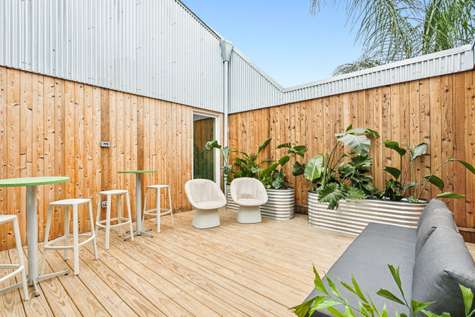 Bright modern outdoor patio with light wood decking and vertical timber fence, corrugated metal wall, two cream lounge chairs, gray sofa, high bar tables with white stools, and corrugated metal planters filled with tropical greenery under a clear blue sky.