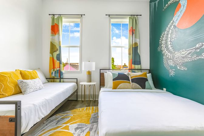 Sunny colorful guest bedroom with two twin beds, yellow and teal pillows, abstract patterned curtains, round side table lamp, yellow-gray rug, and a teal accent wall with a playful white crocodile mural.