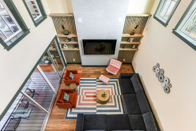 Aerial view of a bright loft-style living room with high ceilings and tall windows, featuring a wall-mounted TV, dark sectional sofa, geometric rug, two rust-orange armchairs, a pink accent chair, round coffee table, hardwood floors and sliding glass doors to a deck.