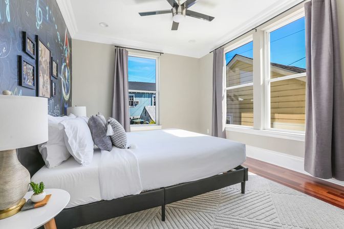 Bright modern bedroom with white bedding and gray pillows, large sunlit windows with gray curtains showing neighboring houses, ceiling fan, hardwood floors and geometric rug.