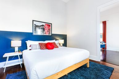 Bright modern bedroom with navy half-wall, wooden platform bed dressed in white linens, red accent pillows, bedside lamps, blue shag rug and hardwood floors, doorway opening to an adjacent sunlit room.