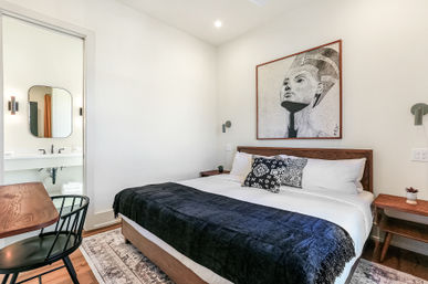 Modern downtown guest bedroom with king bed, wooden headboard, black throw blanket and patterned pillows, framed Egyptian-style portrait above the bed, bedside tables and wall sconces, small wooden desk and chair, and an en-suite bathroom sink and mirror visible — neutral minimalist decor.