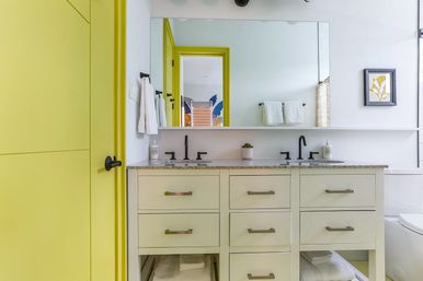 Cheery modern bathroom with a yellow door and frame, large mirror above a double-sink cream vanity with granite countertop, matte black faucets, white towels and a small potted succulent.