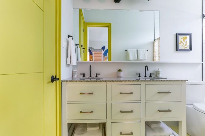 Cheery modern bathroom with a yellow door and frame, large mirror above a double-sink cream vanity with granite countertop, matte black faucets, white towels and a small potted succulent.