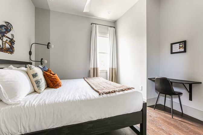 Bright, modern small urban apartment bedroom—white bedding with orange accent pillows, black wall sconce, curtained window, and compact wall-mounted desk.