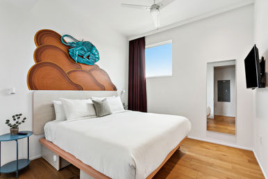 Bright modern minimalist bedroom with king bed and white linens, hardwood floors, turquoise stingray wall sculpture over a wood-pattern headboard, small round side table, window with burgundy curtain, wall-mounted TV and full-length mirror.