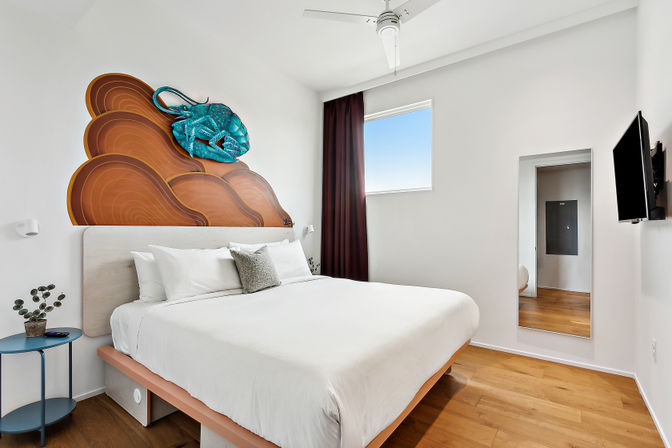 Bright modern minimalist bedroom with king bed and white linens, hardwood floors, turquoise stingray wall sculpture over a wood-pattern headboard, small round side table, window with burgundy curtain, wall-mounted TV and full-length mirror.