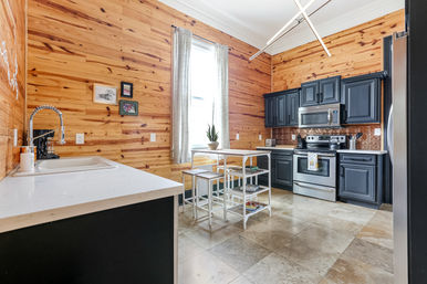 Bright wood-paneled kitchen with navy cabinets and stainless steel appliances, white quartz counters, small white island with stools beneath a tall window, tile floor, modern farmhouse vibe.