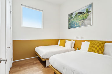 Bright minimalist guest bedroom with two white beds, mustard-yellow accent stripe and matching textured pillows, hardwood floors, small high window and framed garden artwork