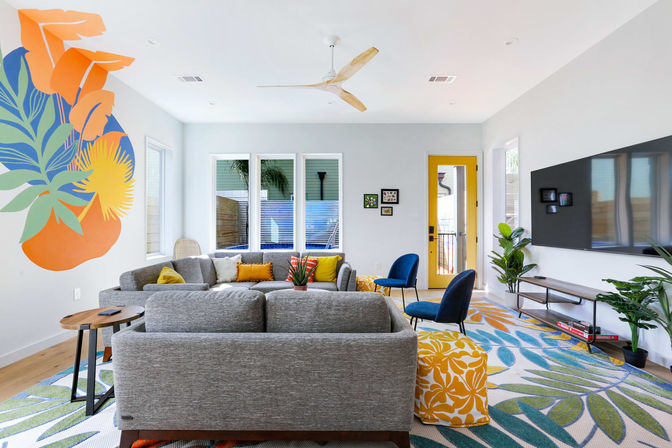 Sunlit modern living room with gray sectional, bold tropical wall mural, blue accent chairs, yellow entry door, and leaf-pattern rug.