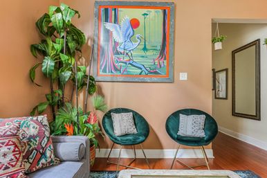 Cozy eclectic living room corner with peach wall, colorful heron painting, two green velvet round chairs with patterned pillows, large potted plants and hardwood floor.