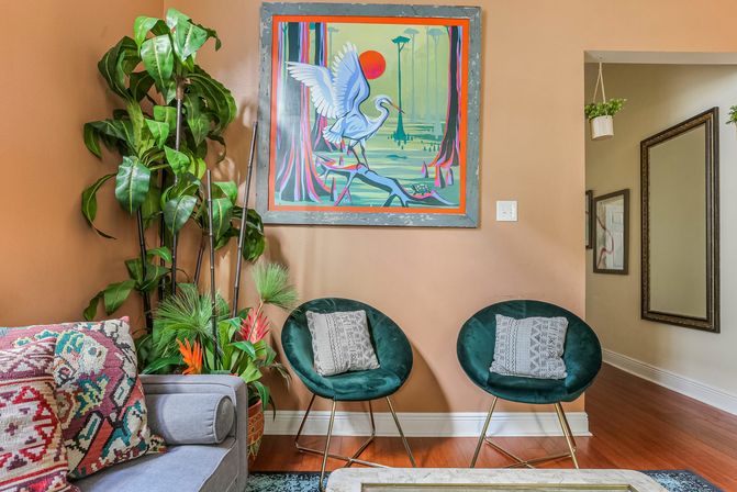 Cozy eclectic living room corner with peach wall, colorful heron painting, two green velvet round chairs with patterned pillows, large potted plants and hardwood floor.