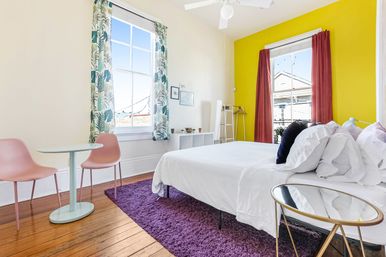 Sunny modern bedroom with a yellow accent wall, two tall windows with tropical-print curtains, white bed on a purple shag rug, hardwood floors, small round table and pink chairs, abundant natural light.
