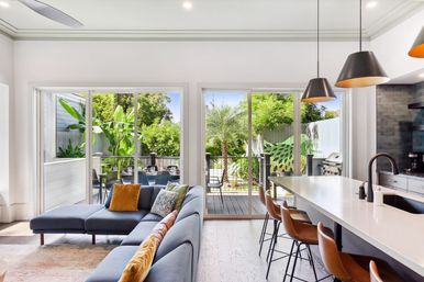 Bright open-plan living room and kitchen with blue sectional and colorful pillows, white island with leather bar stools and pendant lights, sliding glass doors to a sunlit deck, outdoor dining area, grill, and lush tropical backyard with banana and palm trees.
