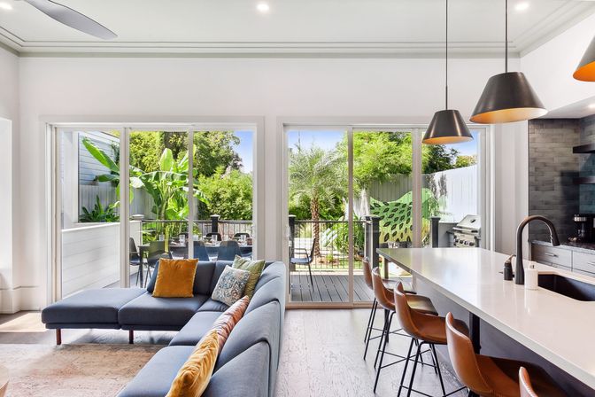 Bright open-plan living room and kitchen with blue sectional and colorful pillows, white island with leather bar stools and pendant lights, sliding glass doors to a sunlit deck, outdoor dining area, grill, and lush tropical backyard with banana and palm trees.