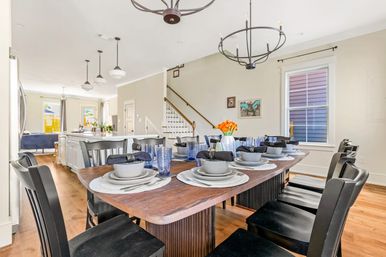 Sunlit open-plan dining room with a long wooden table set for eight—black chairs, blue glassware, gray place settings and an orange flower centerpiece—chandelier and pendant lights, hardwood floors, with a view into the white kitchen and staircase.