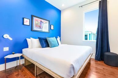 Bright modern bedroom with cobalt blue accent wall, white platform bed with navy and teal pillows, framed artwork, marble side table, hardwood floors and a window with dark curtain letting in daylight.