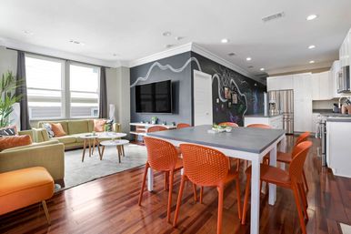 Bright modern open-plan living room and kitchen in an urban apartment with hardwood floors, green sofa, orange dining chairs, gray dining table, chalkboard accent wall with wall-mounted TV, large windows and stainless-steel appliances.