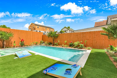 Urban backyard with rectangular swimming pool, wooden privacy fence, artificial turf, cornhole boards and tropical plants under a bright blue sky
