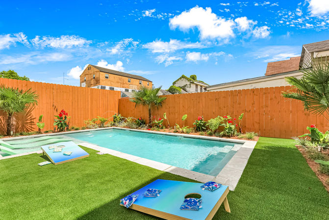 Urban backyard with rectangular swimming pool, wooden privacy fence, artificial turf, cornhole boards and tropical plants under a bright blue sky