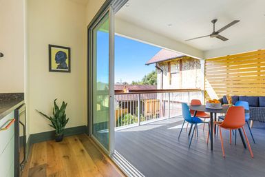 Sunny covered balcony with sliding glass door opening to a modern outdoor dining area: round table with blue and orange chairs, ceiling fan, wicker sofa, wooden slat privacy screen, potted plant and suburban rooftops under clear blue sky.