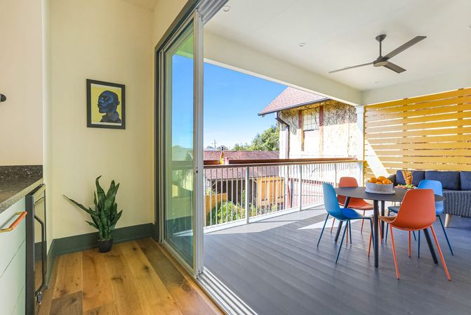 Sunny covered balcony with sliding glass door opening to a modern outdoor dining area: round table with blue and orange chairs, ceiling fan, wicker sofa, wooden slat privacy screen, potted plant and suburban rooftops under clear blue sky.