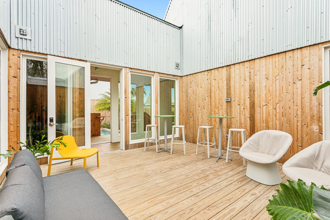 Sunny modern wooden courtyard patio with light wood deck and corrugated metal upper walls, sliding glass doors to the interior, gray sofa and yellow lounge chair, two white swivel chairs, high-top tables with stools, and potted tropical plants.