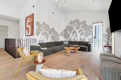 Bright modern living room with vaulted ceiling and pale wood floors, charcoal L-shaped sectional beneath a gray tree wall mural, round wooden coffee table on a braided rug, mustard-yellow lounge chairs and a large wall-mounted TV.