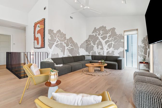 Bright modern living room with vaulted ceiling and pale wood floors, charcoal L-shaped sectional beneath a gray tree wall mural, round wooden coffee table on a braided rug, mustard-yellow lounge chairs and a large wall-mounted TV.