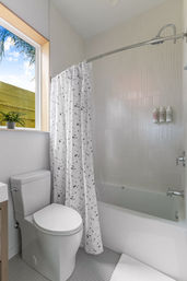 Bright modern small bathroom with white tub-shower and textured wall tiles, curved shower rod with terrazzo-pattern curtain, compact white toilet and sunny window with a potted plant.