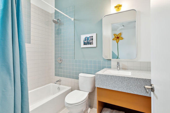 Modern bathroom with tub-shower combo, pale blue square tile accent wall and blue shower curtain, white toilet, speckled gray vanity countertop over orange cabinet, rectangular mirror and yellow flower wall decor