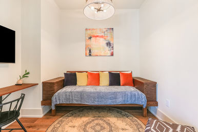 Cozy modern home interior: wooden daybed with blue throw and colorful orange, yellow and charcoal pillows beneath abstract wall art, round patterned rug, pendant light and small floating desk with plant.
