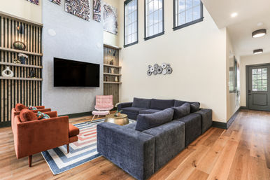 Sunlit modern living room with high ceilings: large dark gray sectional, two rust-colored velvet armchairs, wall-mounted flat-screen TV, geometric area rug, built-in wood shelving with decor, tall windows and warm hardwood floors.
