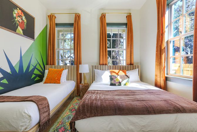 Sunlit, vibrant guest bedroom with a twin bed and a larger bed, striped headboards, bold green geometric mural, orange curtains, floral accent pillows and a colorful runner rug, windows looking out to tree-lined yard.