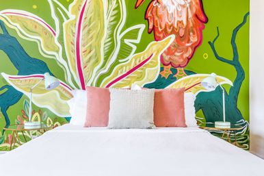Bright tropical bedroom with white bed, three coral and pale-gray pillows, mint bedside lamps and a vivid lime-green mural of oversized leaves and a colorful bird