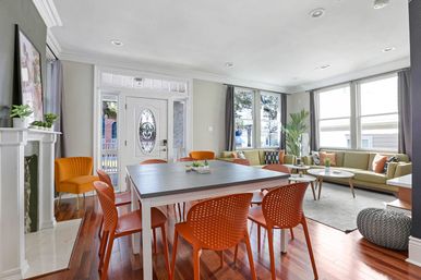 Sunlit open-plan living and dining room with decorative front door, gray dining table surrounded by orange modern chairs, olive-green sectional sofa, hardwood floors, tall windows and indoor plants.