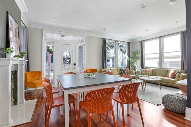 Sunlit open-plan living and dining room with decorative front door, gray dining table surrounded by orange modern chairs, olive-green sectional sofa, hardwood floors, tall windows and indoor plants.