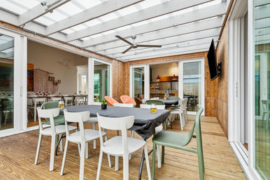 Covered wood-paneled sunroom dining area with mixed modern chairs around black-clothed tables, two orange lounge chairs, ceiling fan, wall-mounted TV, and sliding glass doors to the kitchen.