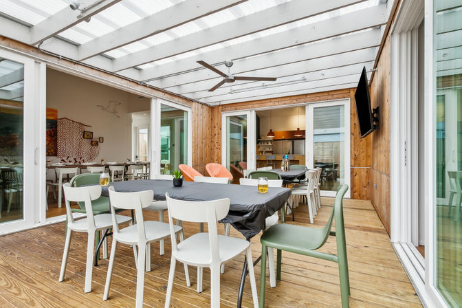 Covered wood-paneled sunroom dining area with mixed modern chairs around black-clothed tables, two orange lounge chairs, ceiling fan, wall-mounted TV, and sliding glass doors to the kitchen.