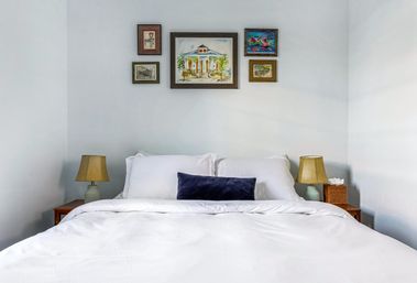 Bright minimalist bedroom interior with white bedding and navy lumbar pillow, framed artwork above the bed, wooden nightstands with matching table lamps and a tissue box