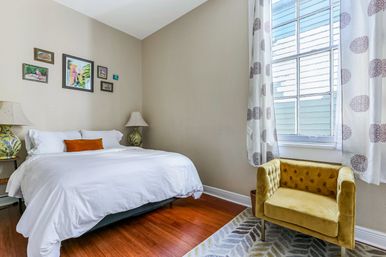 Cozy bright bedroom with white bedding and orange lumbar pillow on a queen bed, framed artwork above the headboard, matching table lamps, warm hardwood floors, large window with patterned white curtains, and a mustard-yellow velvet armchair on a patterned rug.