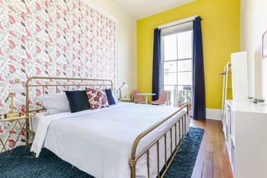 Cheerful modern bedroom featuring a brass bed with white linens, pink bird-pattern wallpaper on one wall and a bold yellow accent wall with navy curtains, small balcony, pink bistro chairs, blue shag rug and brass bedside lamps.