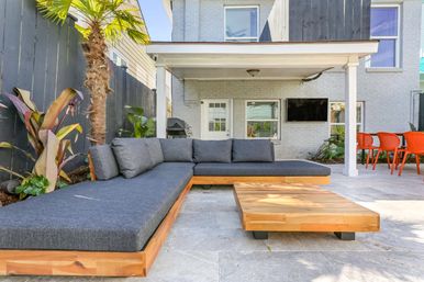 Sunny backyard patio with modern L-shaped wooden sectional topped with dark gray cushions, low wooden coffee table, palm and tropical plants, covered porch with wall-mounted TV and grill, and bright orange outdoor dining chairs.